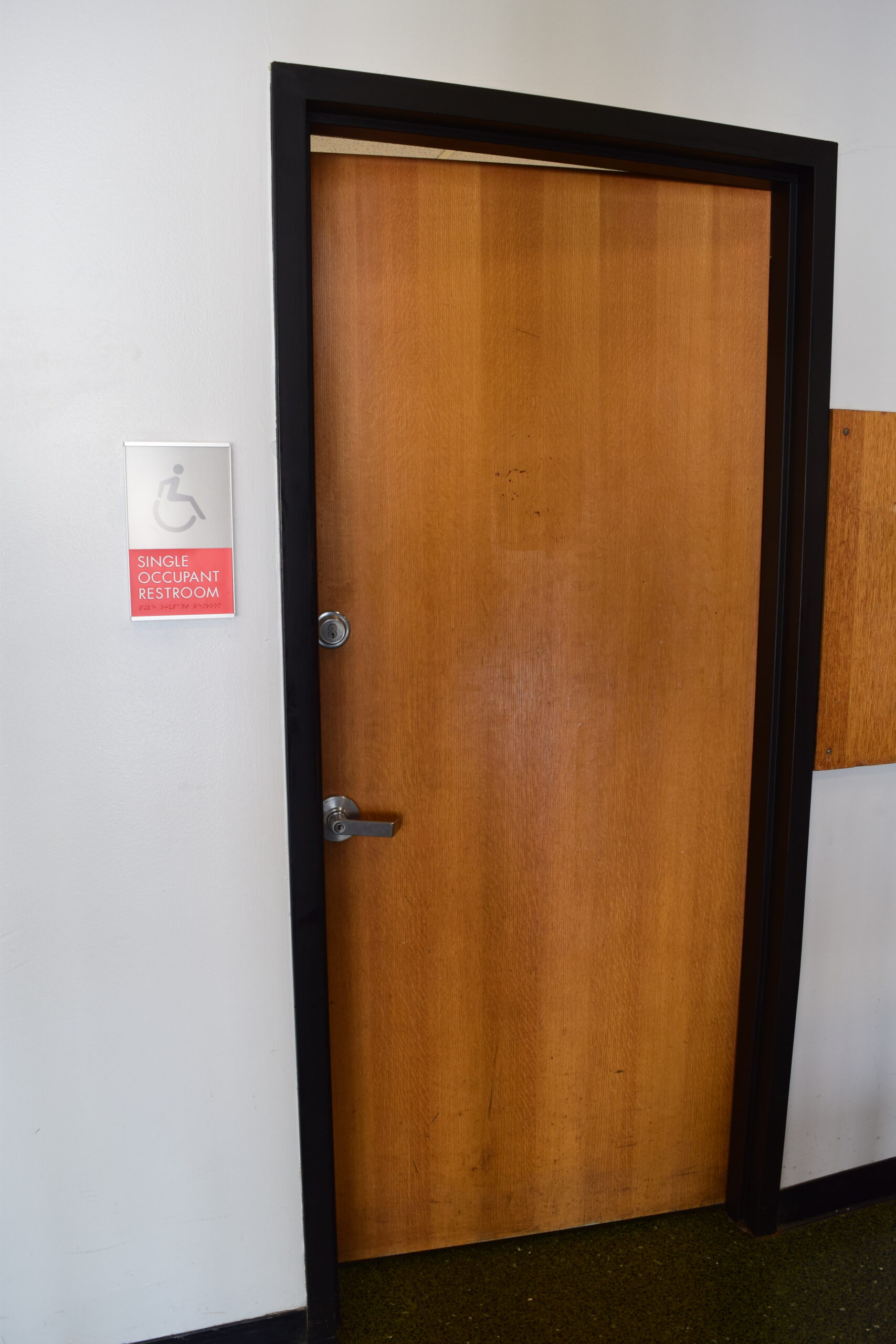 A wooden door opens inward to a single-use bathroom, with a lever handle low on the left of the door, and a deadbolt lock above it. A sign next to the door has an accessibility icon and says "Single occupant restroom."