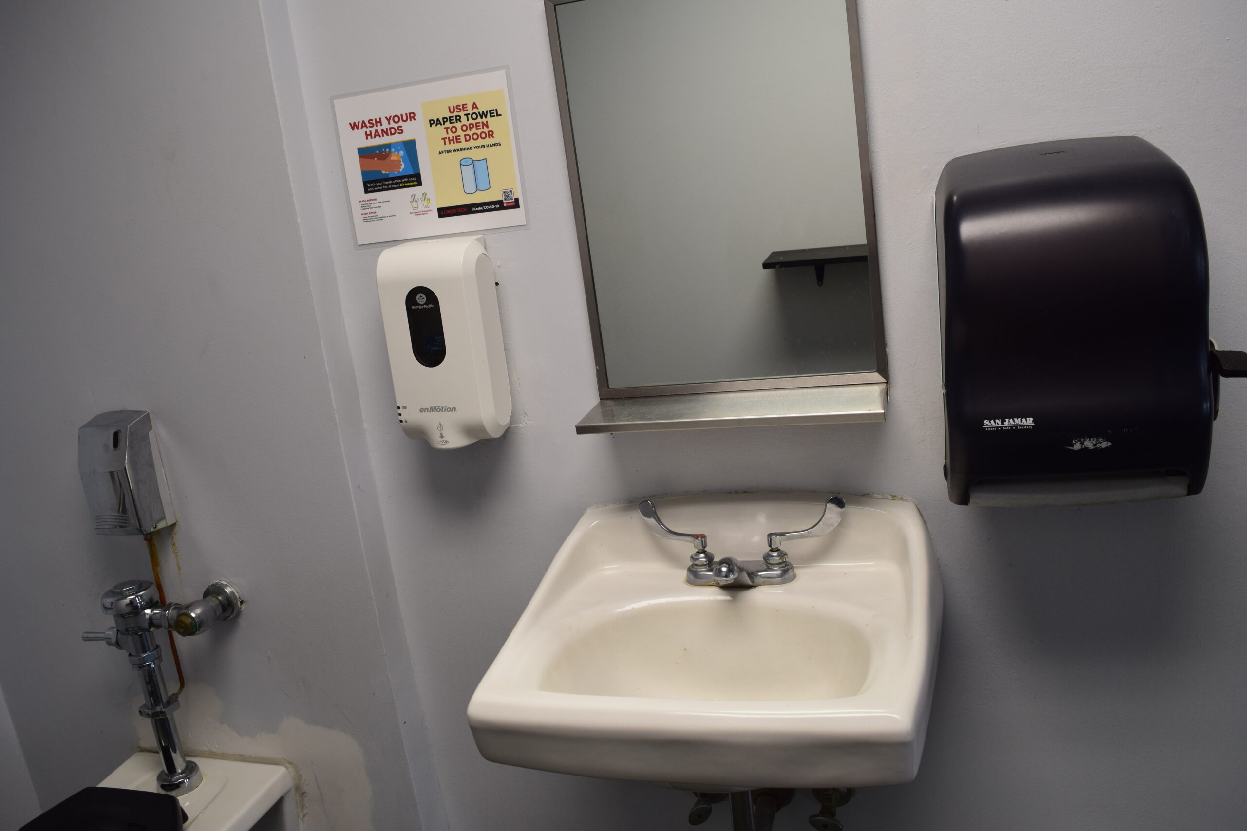 A sink with large hot and cold knobs, with an automatic soap dispenser on the left and a manually-operated paper towel dispenser on the right.