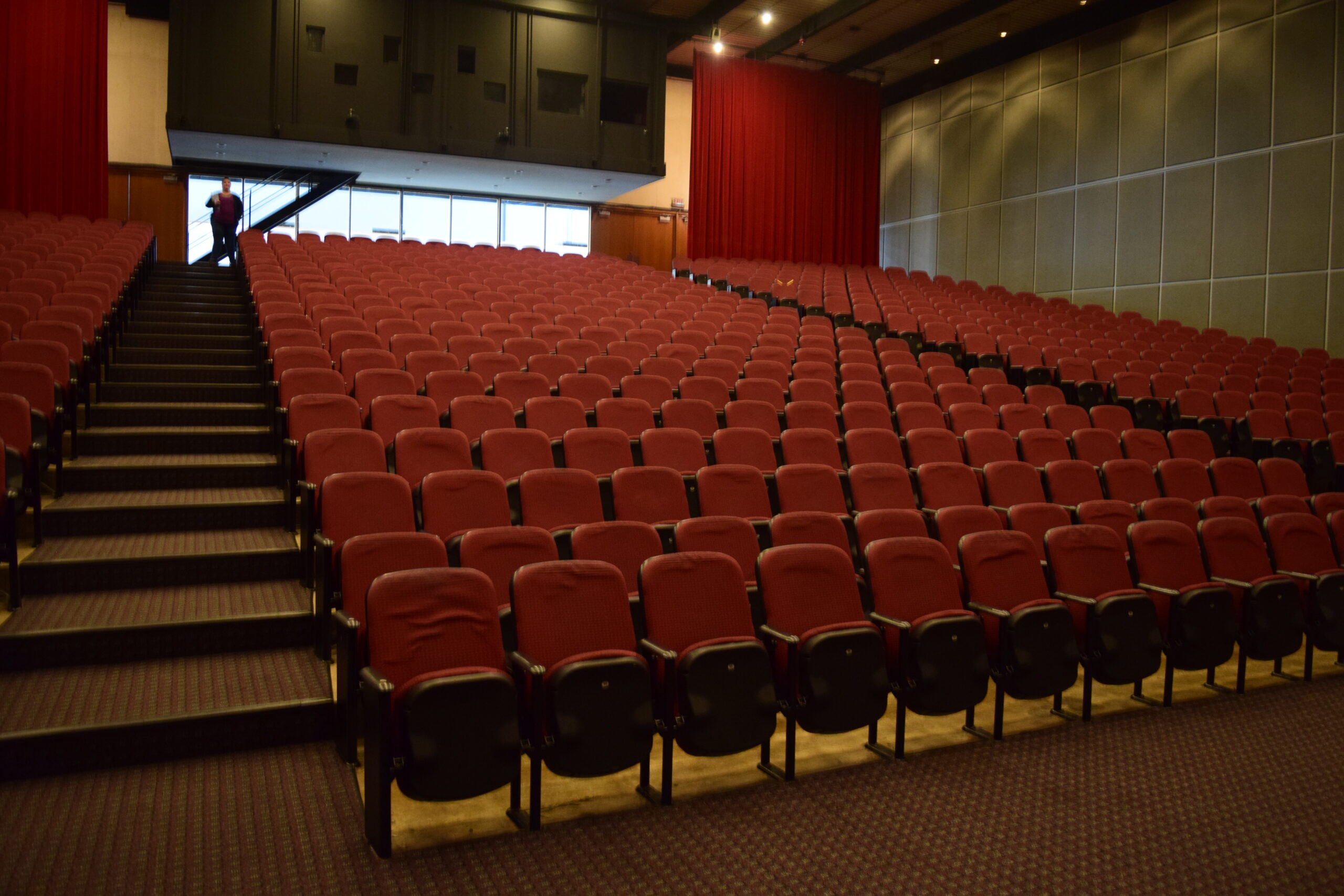 A large auditorium seen from the front, with two aisles ascending in long steps, and three blocks of red cushioned theater chairs.