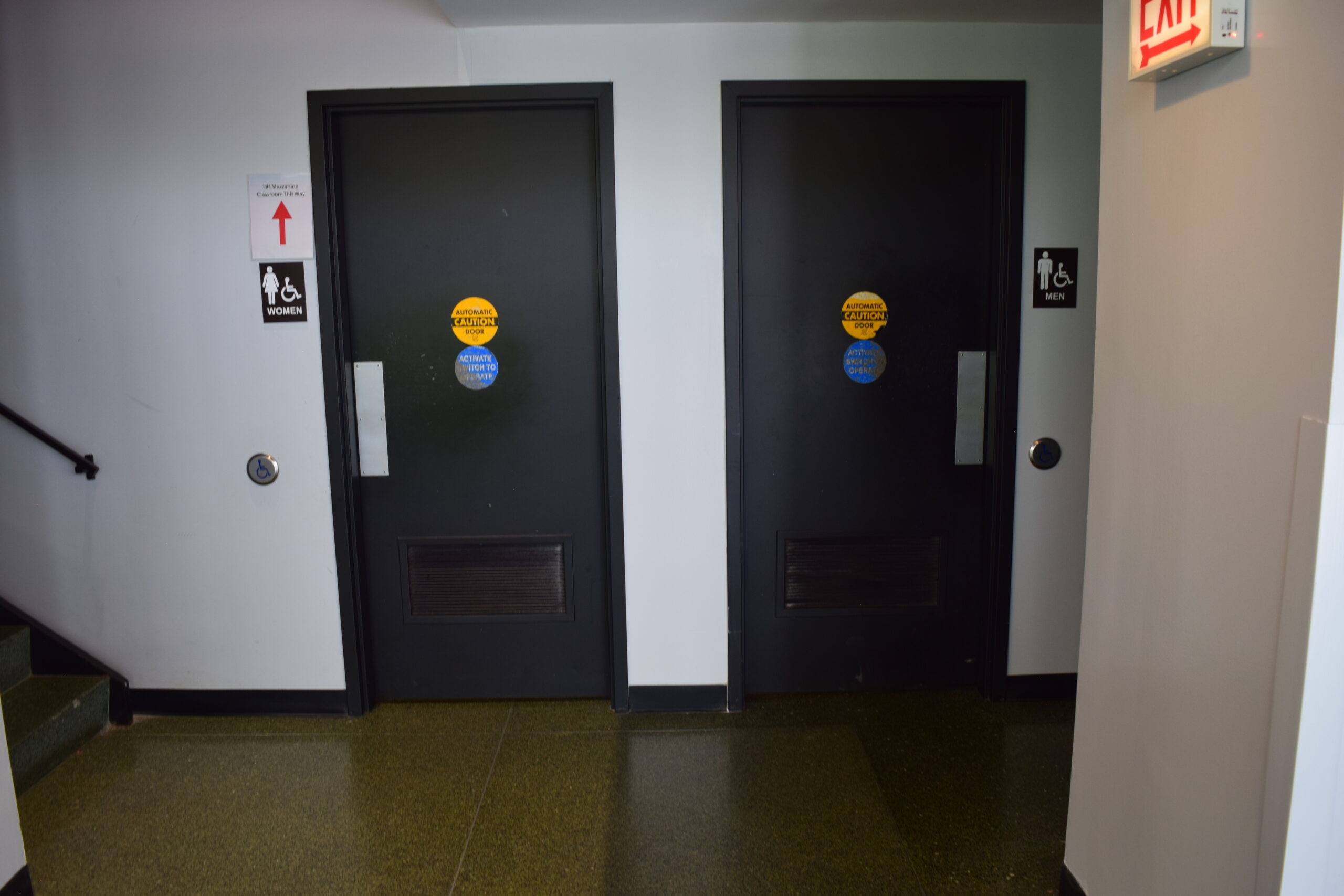A pair of doors with push plates. The left door has a sign that says "Women" and has female and accessibility icons. The right door has a sign that says "Men" and has male and accessibility icons.