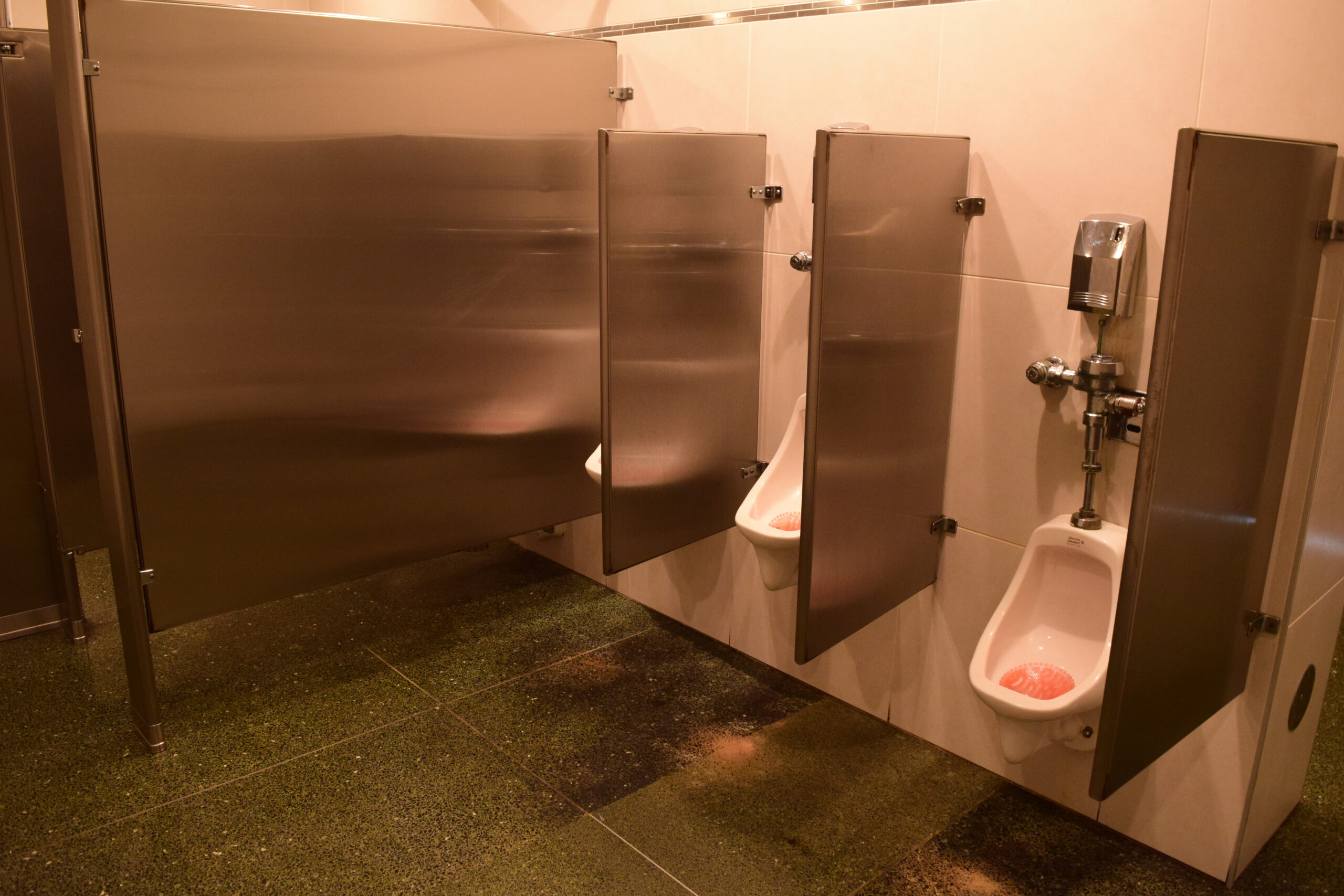 A set of three urinals separated by metal dividers. The urinal on the right is lower to the ground.