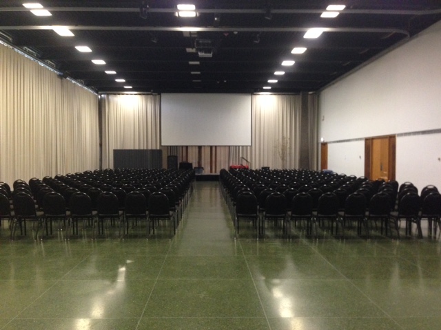 A large room set up conference-style with a large center aisle, two large blocks of chairs, and a projector screen at the front.